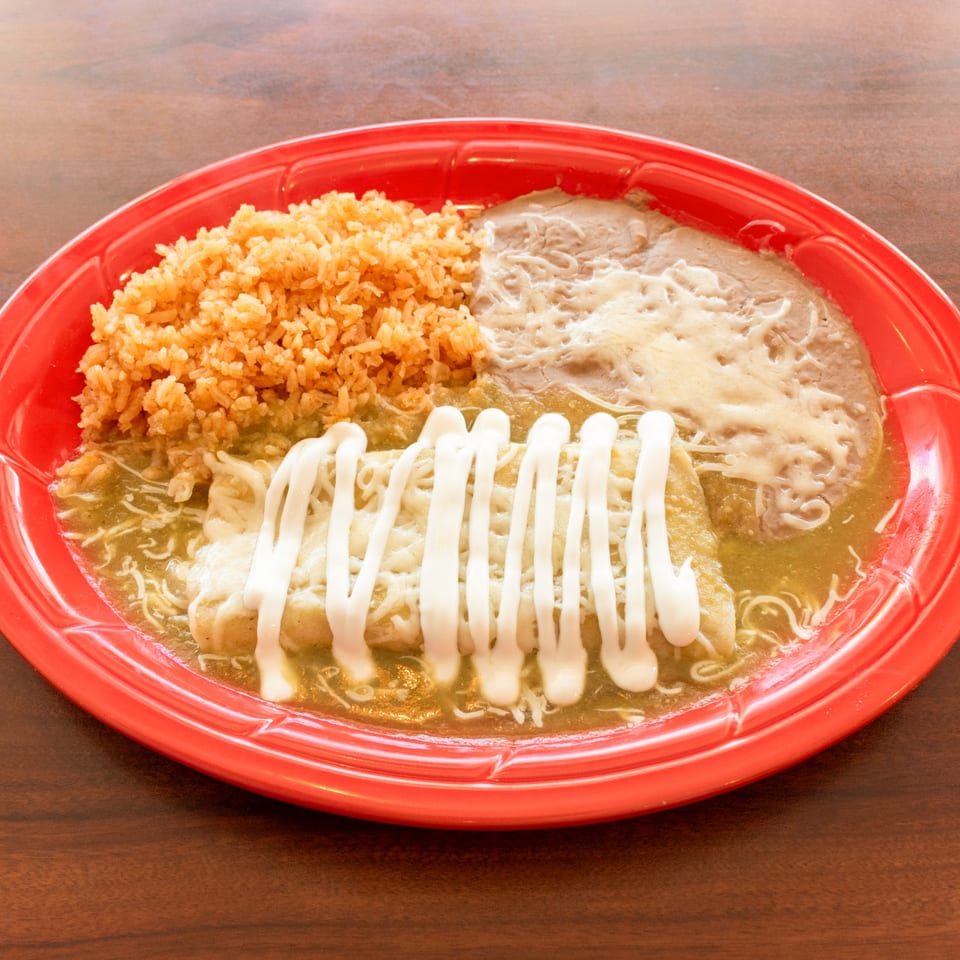 Red plate with enchiladas covered in green sauce and sour cream, rice, and refried beans.