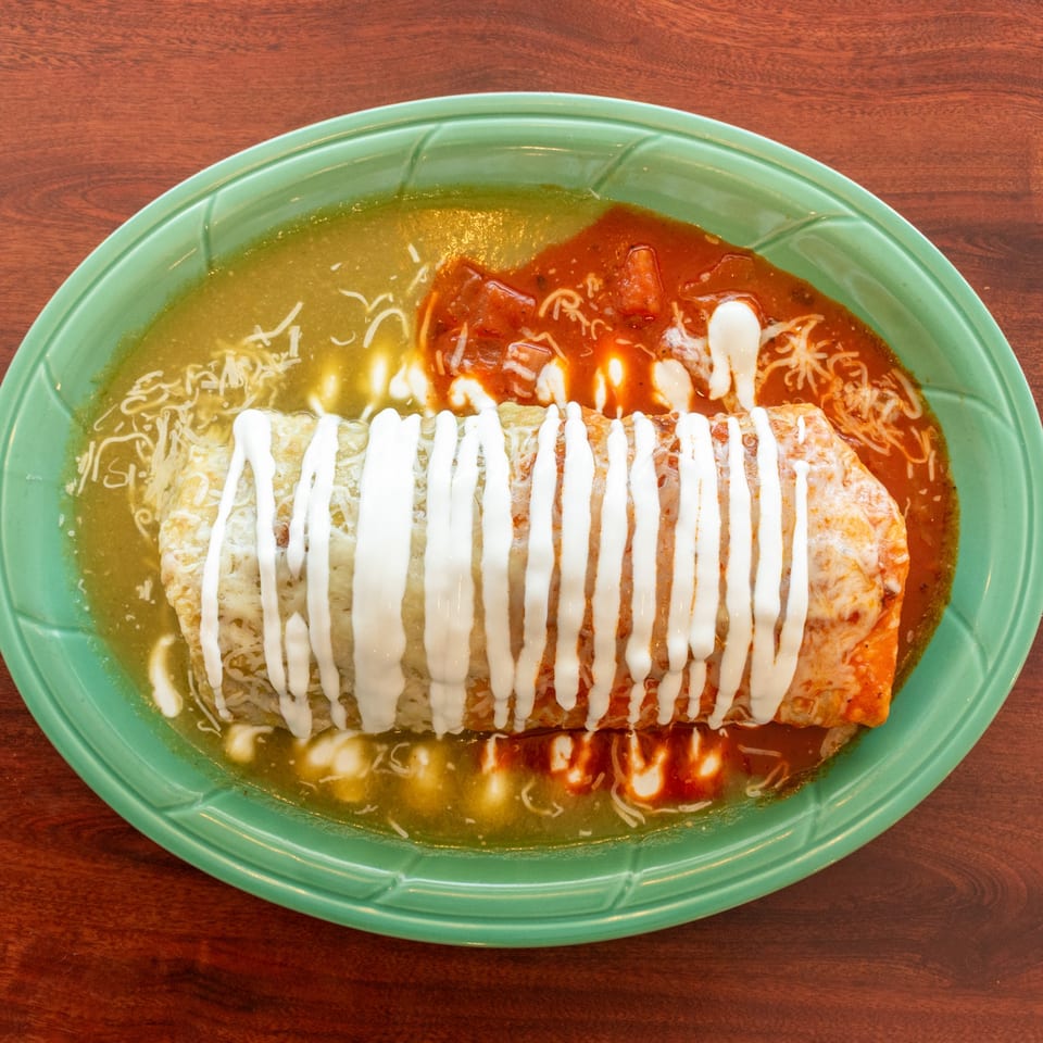 A large burrito covered in green and red sauce, cheese, and sour cream on a green plate.