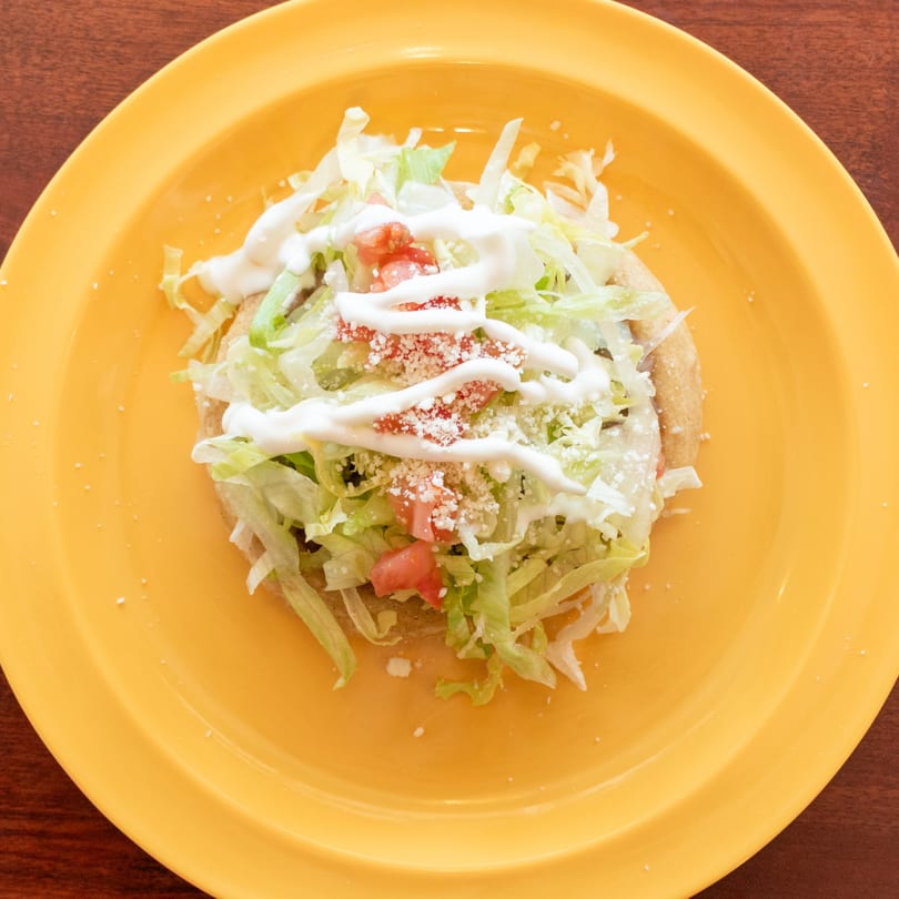 A sopes on a yellow plate, topped with shredded lettuce, diced tomatoes, cheese, and crema.