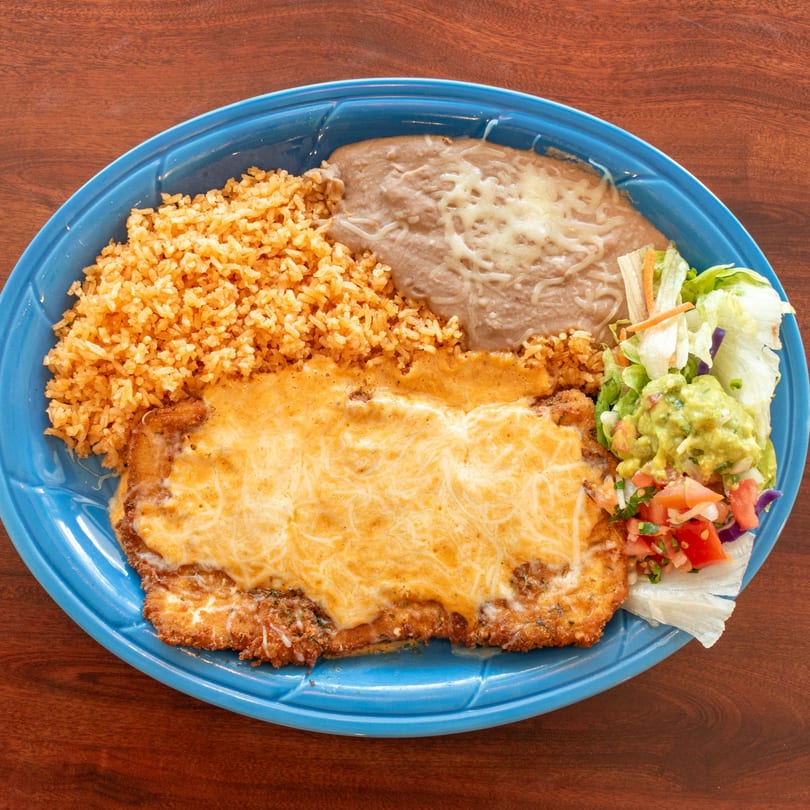 A blue plate holds a cheesy chicken milanesa, rice, refried beans, guacamole, and pico de gallo.