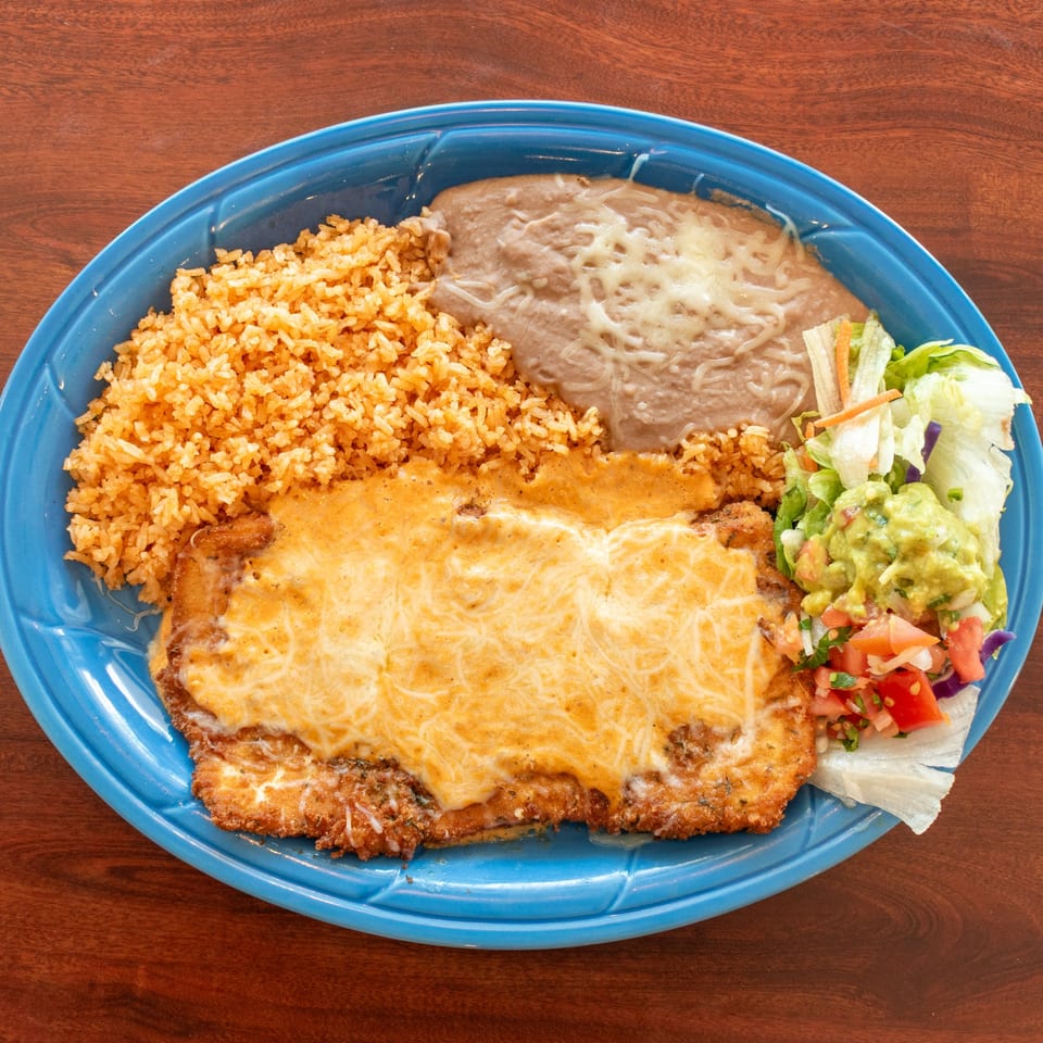 A blue plate holds a cheesy chicken dish with rice, refried beans, guacamole, and pico de gallo.