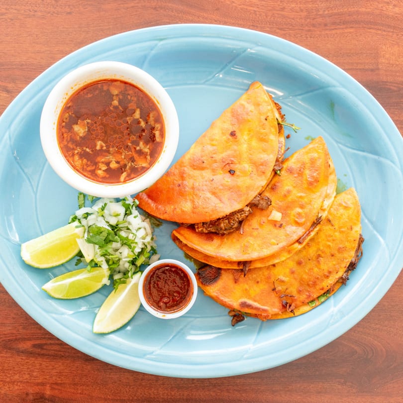 Birria tacos with consommé, salsa, chopped onions, cilantro, and lime wedges on a blue plate.
